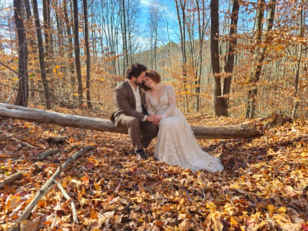 A couple in wedding attire sitting on a fallen log in the autumn forest at Raven's Retreat