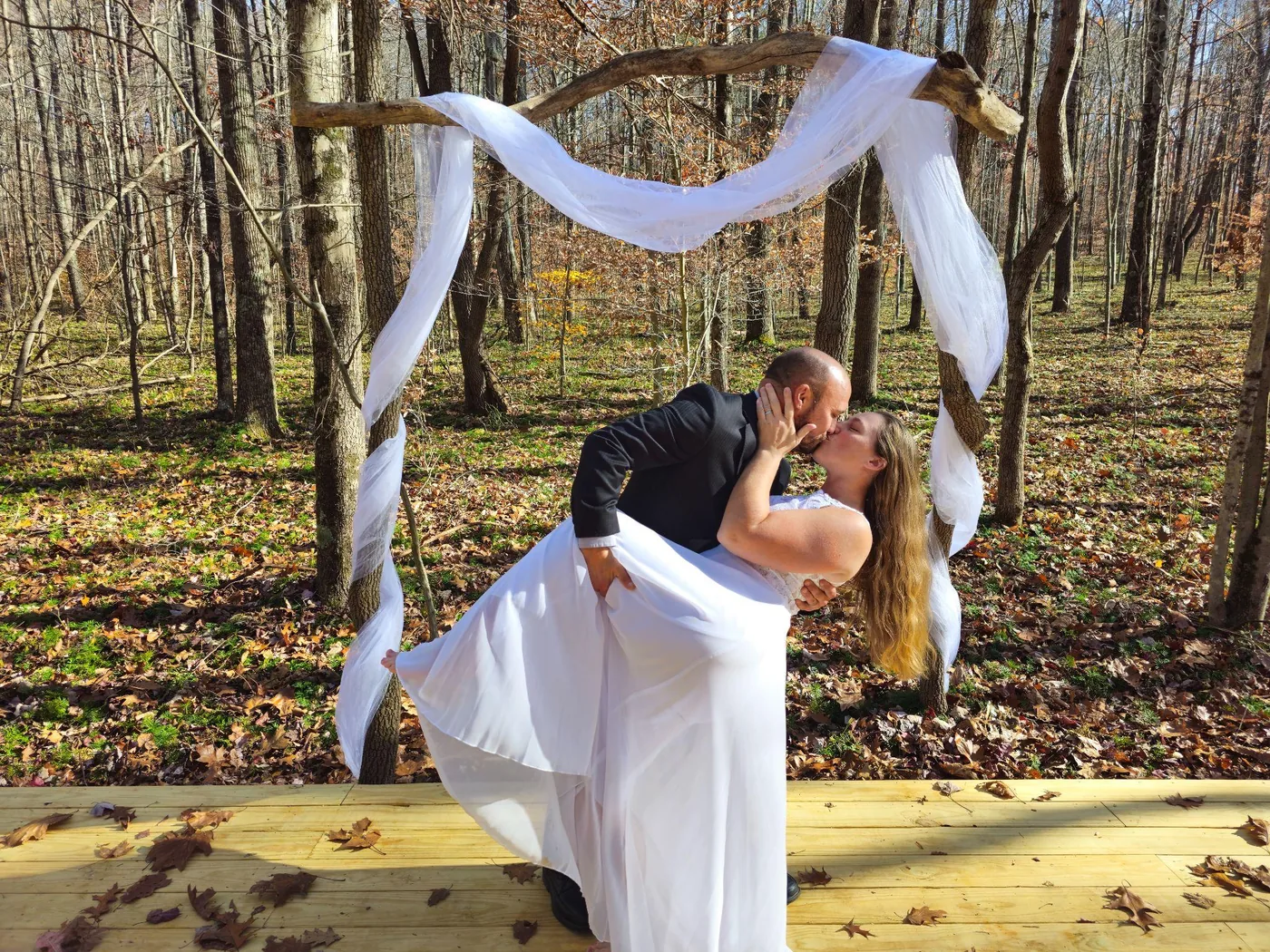 Couple kissing beneath a draped wooden arch on the forest ceremony platform at Raven’s Retreat